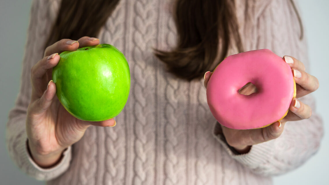 Young woman holding a doughnut and green apple in her hands. Concept of healthy and unhealthy food, clean eating, lifestyle, dieting, and fitness